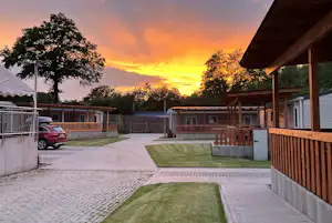 Campingplatz mit gepflastertem Weg, Rasenfläche und Holzunterstand am Rand, im Hintergrund Wohnmobile und Zaun bei Sonnenuntergang.