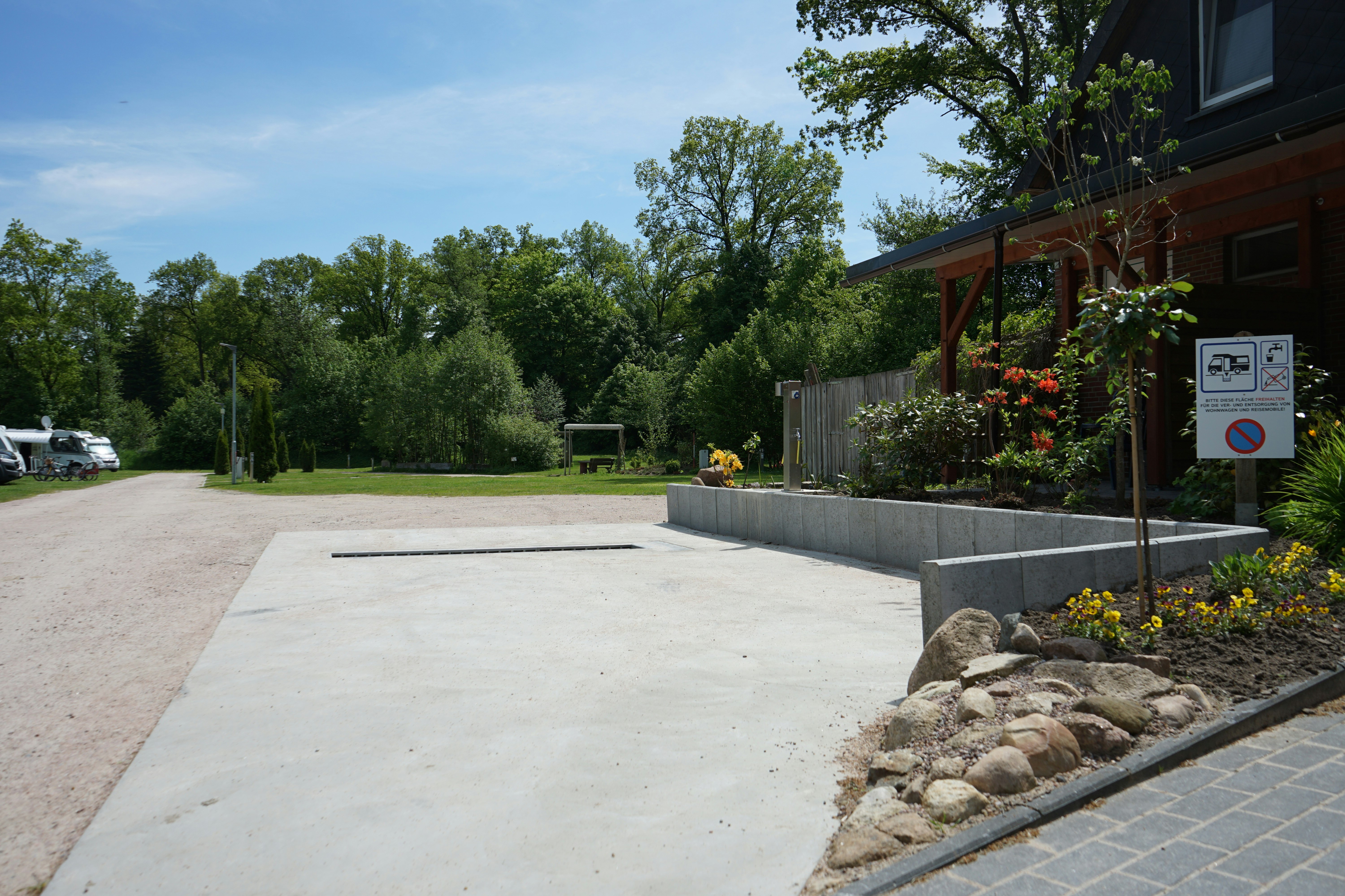 Betonweg und Terrasse am Campingplatz, mit Blumenbeeten, Steinkante und Bäumen im Hintergrund.