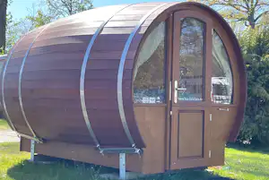 Rundes Fass-Campinghaus mit Holztür und Fenster, auf grünem Rasen im Campinggelände, im Hintergrund Bäume.