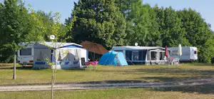 Campingplatz mit mehreren Wohnwagen und Zelten auf einer Wiese, im Hintergrund Bäume und ein weißes Sanitärgebäude.