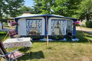 Campingzelt mit Terrasse, Tisch und Stühlen auf einer Rasenfläche, im Hintergrund Bäume und ein weiteres Zelt.