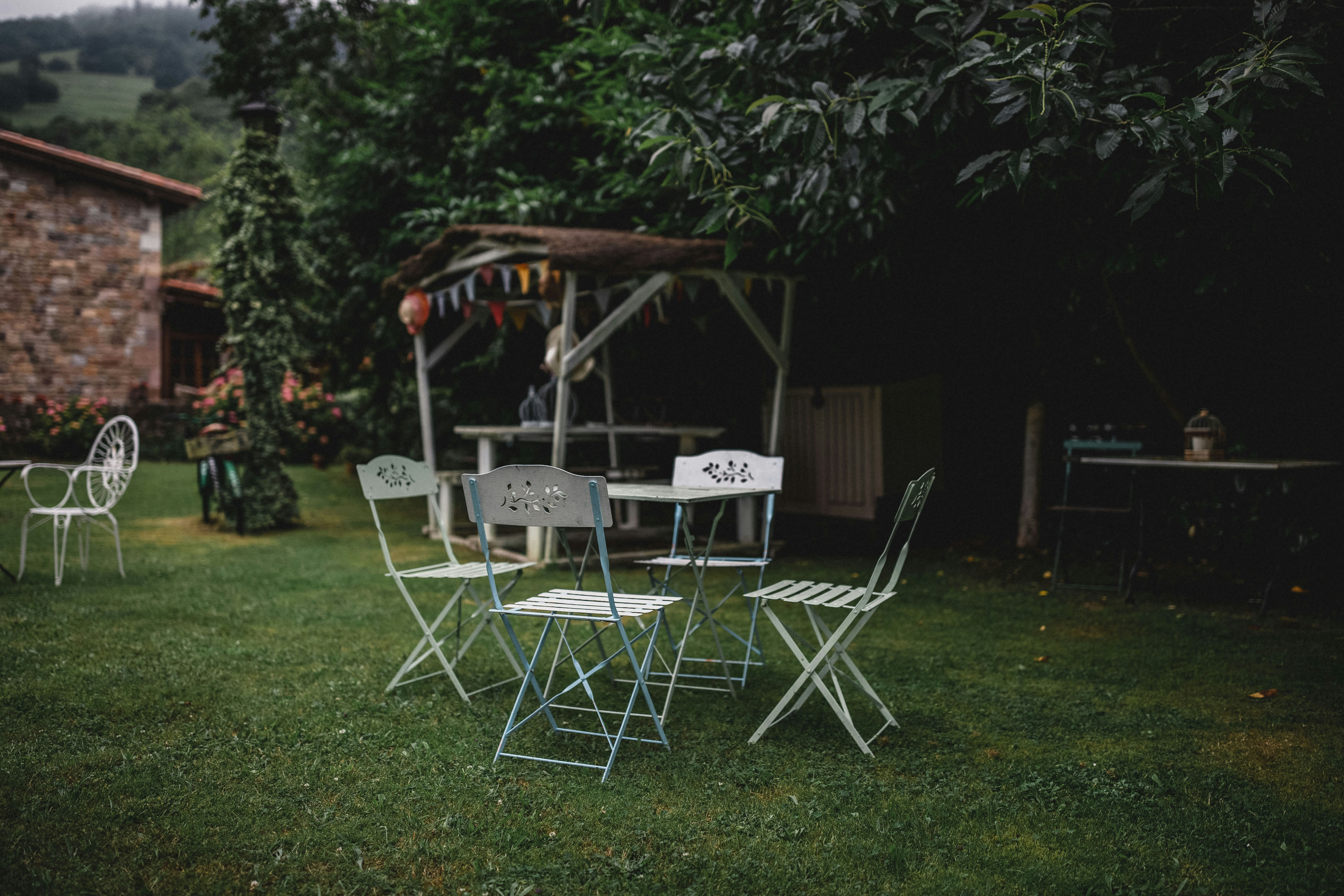 Camping-Esstisch mit Klappstühlen im Garten, neben Steinwand und Rasen, umgeben von Bäumen und Pflanzen.