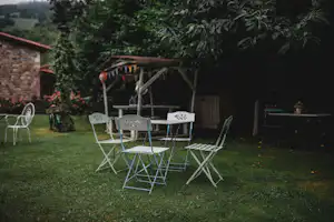 Camping-Esstisch mit Klappstühlen im Garten, neben Steinwand und Rasen, umgeben von Bäumen und Pflanzen.