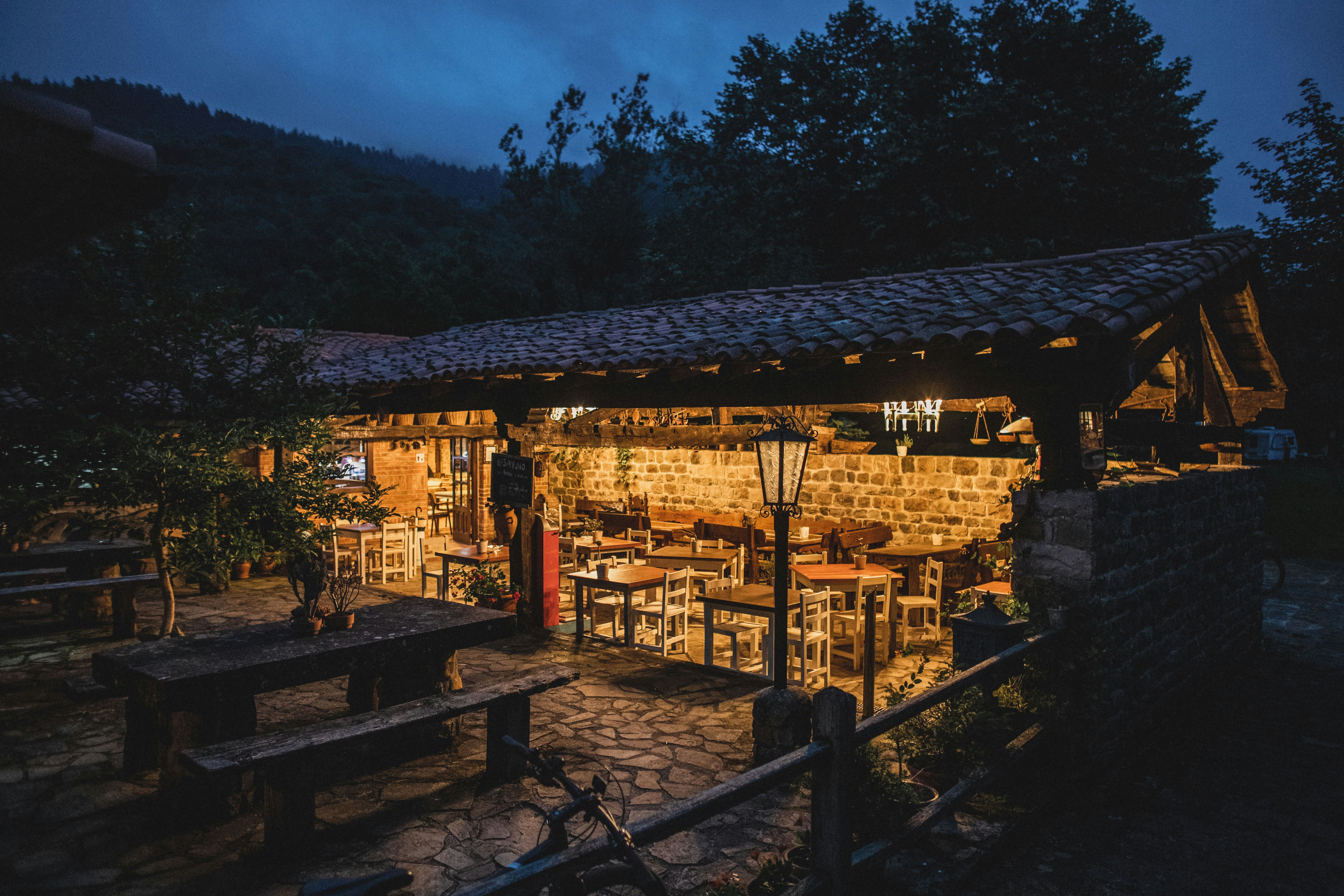 Beleuchtete Terrasse eines Camping-Restaurants mit Holzdach, Tischen und Stühlen, im Hintergrund Bäume und Hügel bei Dämmerung.