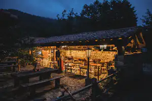 Beleuchtete Terrasse eines Camping-Restaurants mit Holzdach, Tischen und Stühlen, im Hintergrund Bäume und Hügel bei Dämmerung.