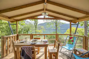 Holzterrasse mit überdachtem Essbereich, Tisch und Stühlen, Blick durch das Geländer auf Bäume und Berge.