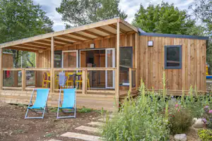 Holzbungalow mit Terrasse, zwei blauen Liegestühlen und umzäuntem Bereich vor dem Gebäude, umgeben von Bäumen und Sträuchern.