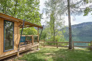 Holzhaus mit verglaster Front und Terrasse, umgeben von Bäumen, mit Blick auf einen See und Hügel im Hintergrund.