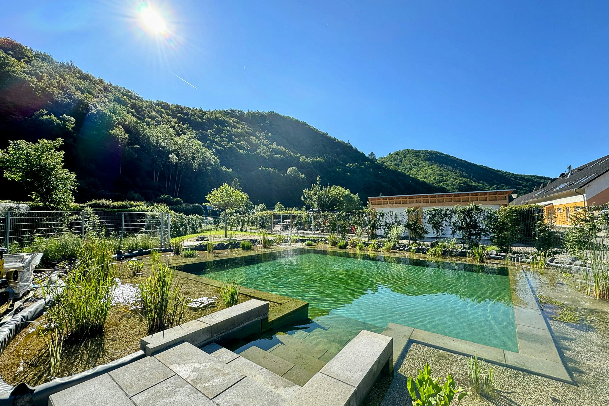 Grüner Camping-Teich mit Steinstufen, Gartenpflanzen und Zaun; im Hintergrund Berge und Bäume unter klarem Himmel.