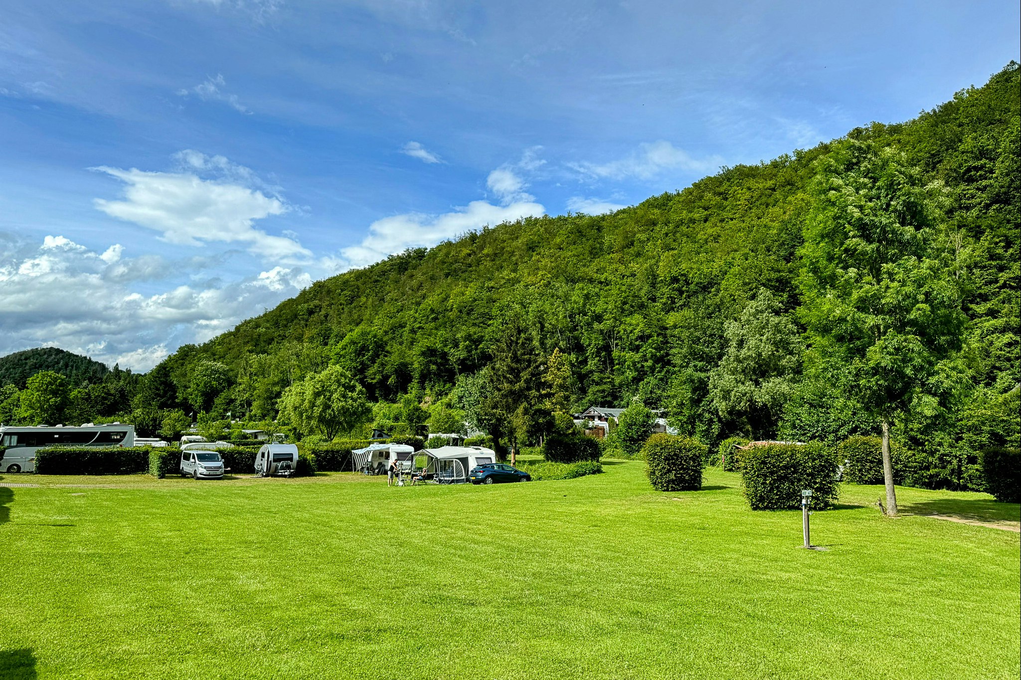 Campingwiese mit Wohnwagen und Zelten, Autos auf dem Stellplatz, umgeben von Hecken und Bäumen unter blauem Himmel.