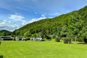 Campingwiese mit Wohnwagen und Zelten, Autos auf dem Stellplatz, umgeben von Hecken und Bäumen unter blauem Himmel.