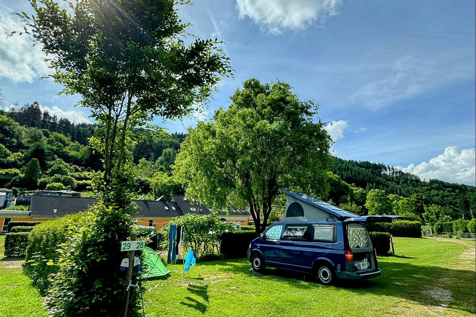Wohnmobil und Zelt auf einem grünen Campingplatz, Hecken und Bäume, daneben ein Parkplatz mit Wohnmobil.