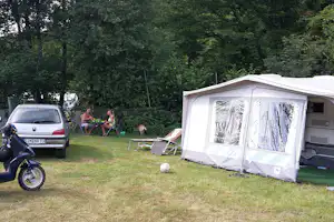 Wohnwagen mit Vorzelt auf Campingwiese, daneben Auto und Motorrad, im Hintergrund Hecke und Bäume.