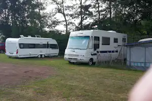 Zwei Wohnmobile auf einem Campingplatz, neben einem Zaun und einem Zelt, umgeben von Bäumen und Rasen.