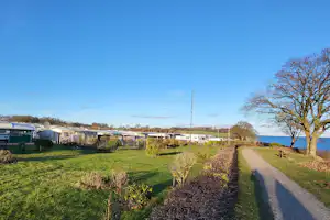 Campingplatz mit gepflegter Rasenfläche, Hecken und Wegen, Wohnwagen und Bungalows am Rand, Blick auf Bäume und Häuser.