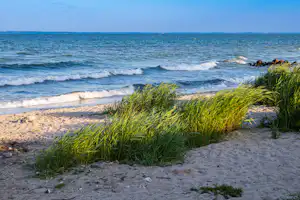 Wellen rollen an den Strand, im Vordergrund Kiesel und Sand, im Hintergrund ruhiges Meer und Horizontlinie.