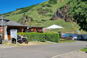 Eingang eines Campingplatzes mit Rezeption, überdachter Terrasse und Fahrrädern, im Hintergrund grüne Hügel und Wiese.