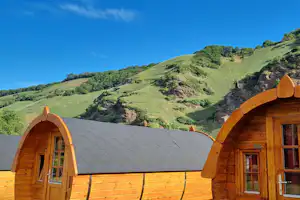 Holz-Glampinghütten mit gewölbten Eingängen auf dem Campingplatz, im Hintergrund grüne Hügel und blauer Himmel.