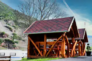 Holzunterstand mit rotem Ziegeldach am Campingplatz, daneben Wohnwagen und Blick auf bewaldete Hänge im Hintergrund.