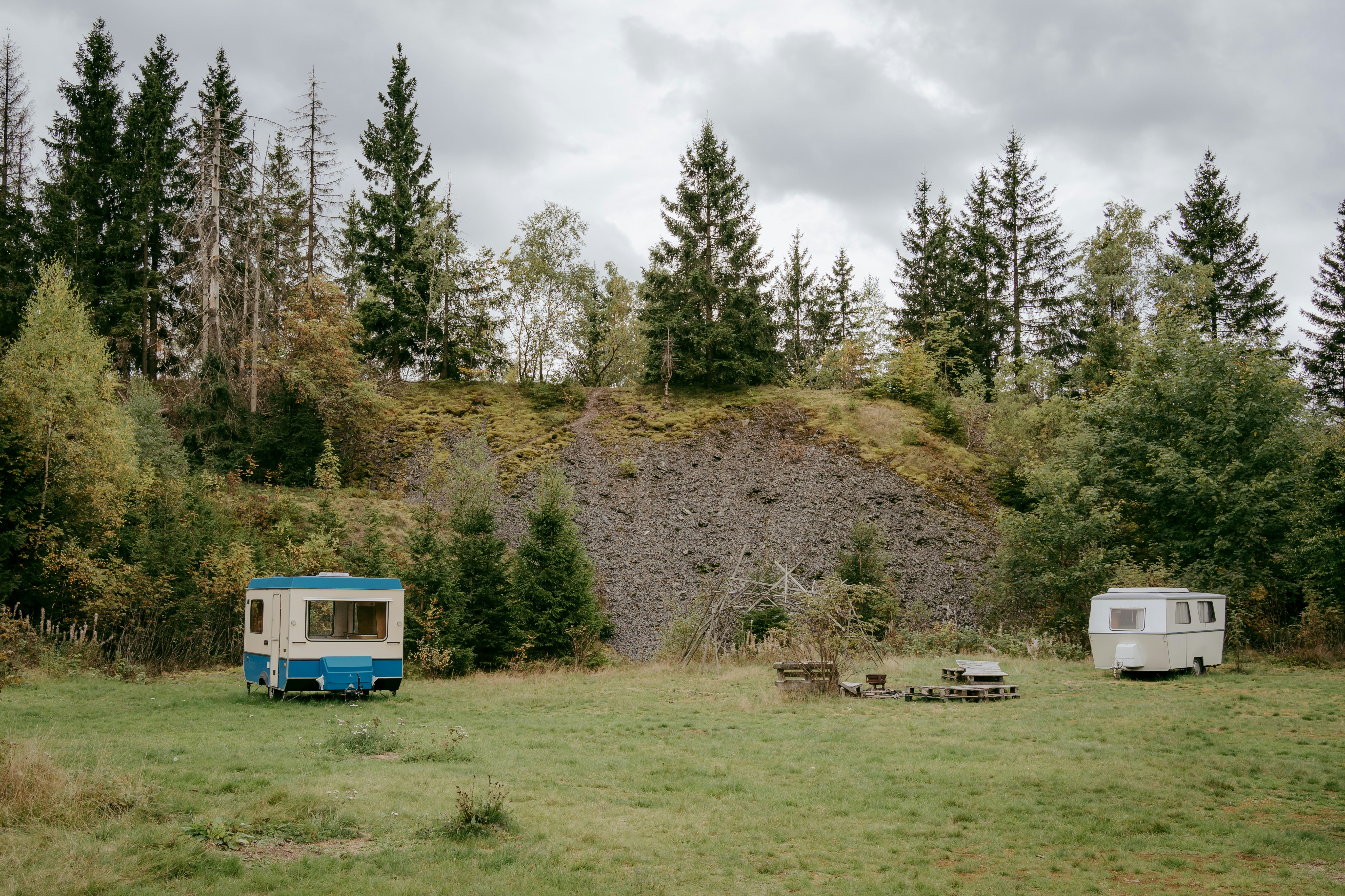 Zwei Wohnwagen auf einer grünen Wiese, mit Sitzbank und Tisch im Vordergrund, umgeben von hohen Bäumen am Hang.