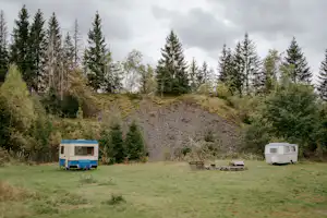 Zwei Wohnwagen auf einer grünen Wiese, mit Sitzbank und Tisch im Vordergrund, umgeben von hohen Bäumen am Hang.