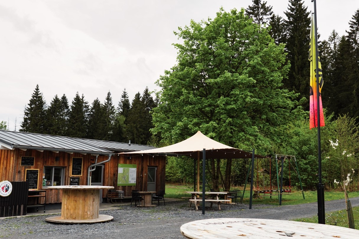 Holzhütten und ein weißes Zeltdach auf dem Campingplatz, mit Sitzbank, Holzsteg und Bäumen im Hintergrund.