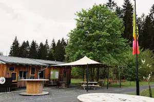 Holzhütten und ein weißes Zeltdach auf dem Campingplatz, mit Sitzbank, Holzsteg und Bäumen im Hintergrund.