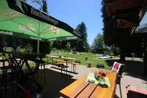 Terrasse mit Holztisch, Blumen und Sitzgelegenheiten, daneben Rasenfläche mit Tischen und Sonnenschirmen am Campingplatz.