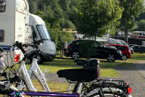 Fahrräder vor einem Wohnmobil auf dem Campingplatz, daneben weitere Autos und Stellplätze mit Bäumen im Hintergrund.