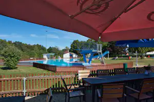 Blick auf den Camping-Pool mit roter Markise, Liegen und Terrasse mit Holztisch und Stühlen im Garten.