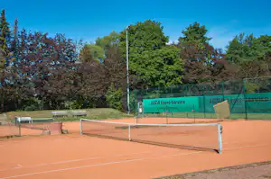 Tennisplatz mit roten Sandbelag, Umzäunung und Netz, im Hintergrund Bäume und ein grünes Schild am Campinggelände.