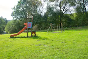 Spielplatz auf einer grünen Wiese: Rutsche, Kletterelement und Schaukel vor Bäumen im Campingbereich.