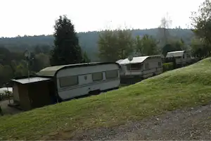 Wohnwagen auf einem Campingplatz am Hang, mit weiteren Fahrzeugen, Bäumen und einem kleinen Gebäude im Hintergrund.