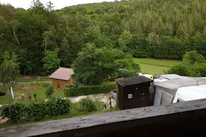Holzhütten und Wohnwagen auf einem Campingplatz, umgeben von Wald und grünen Wiesen, Blick von einer Holzterrasse.