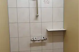 Duscharmatur an einer Wand mit hellen Fliesen, Handbrause und Ablage im kleinen Sanitärbereich.