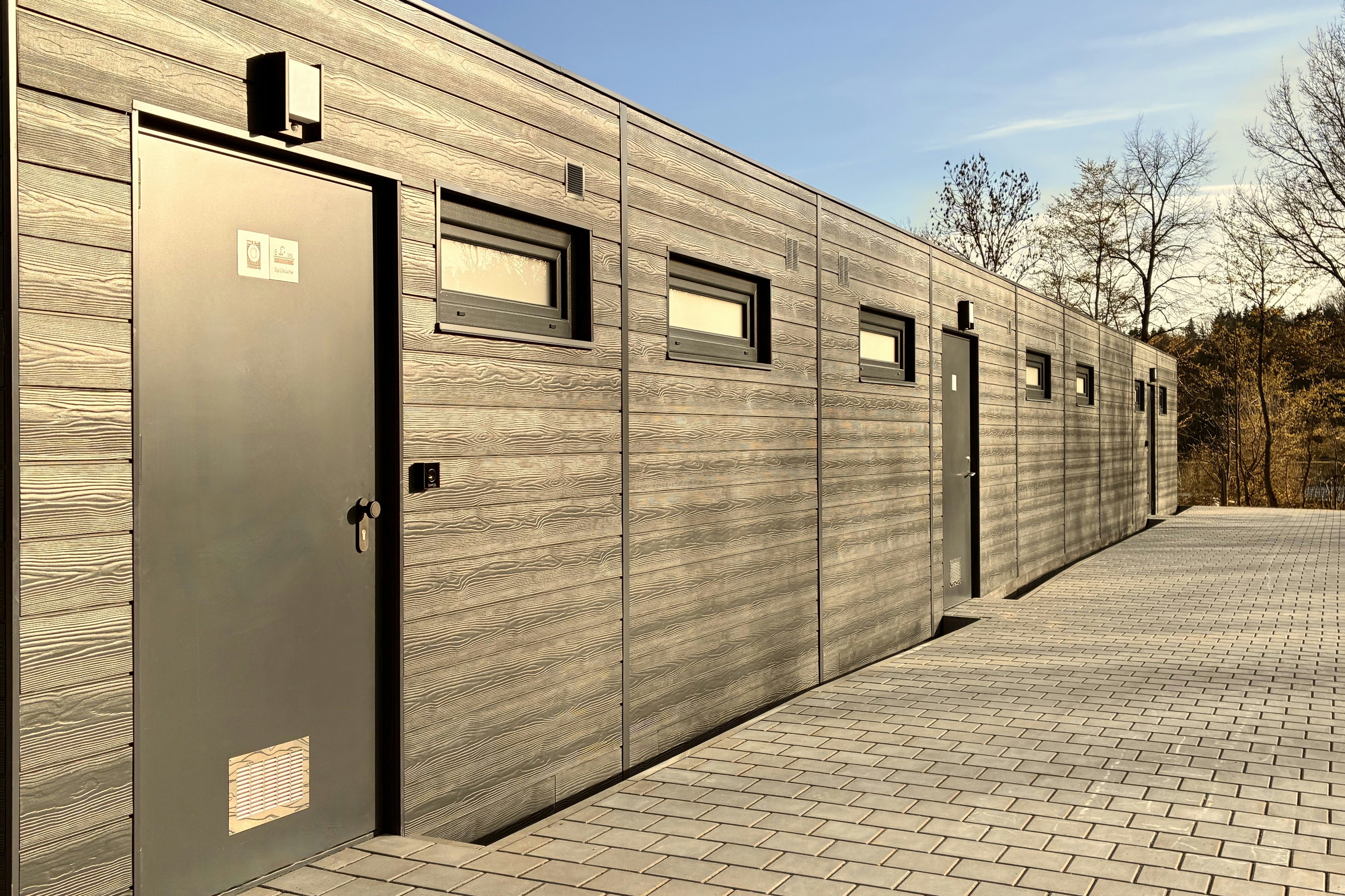 Reihe moderner Holz- und Metallunterkünfte mit Türen und Fenstern, gepflasterter Weg und Bäume am Rand.
