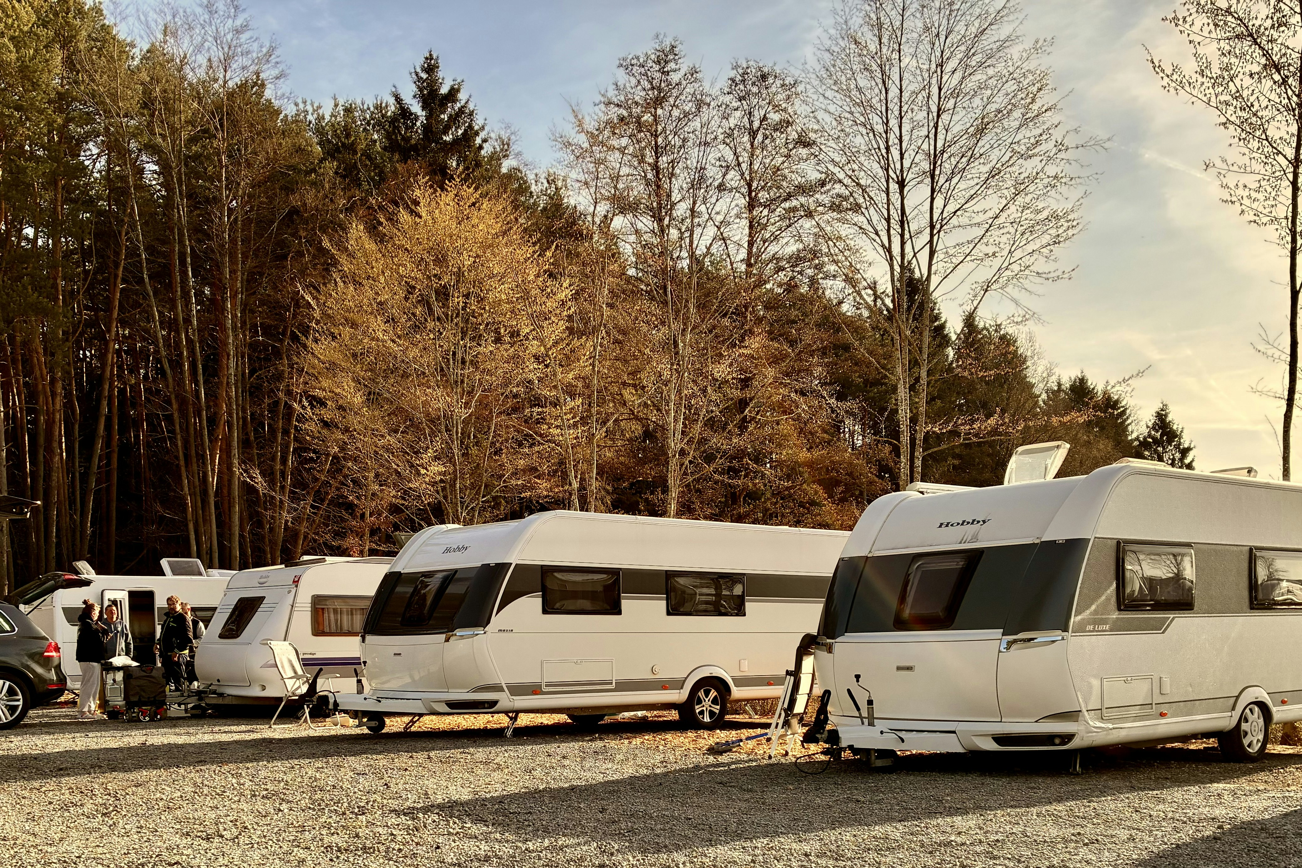 Wohnmobile und Caravans auf einem Campingplatz, geparkte Fahrzeuge am Rand, dahinter Bäume im Herbst.