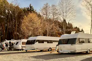 Wohnmobile und Caravans auf einem Campingplatz, geparkte Fahrzeuge am Rand, dahinter Bäume im Herbst.