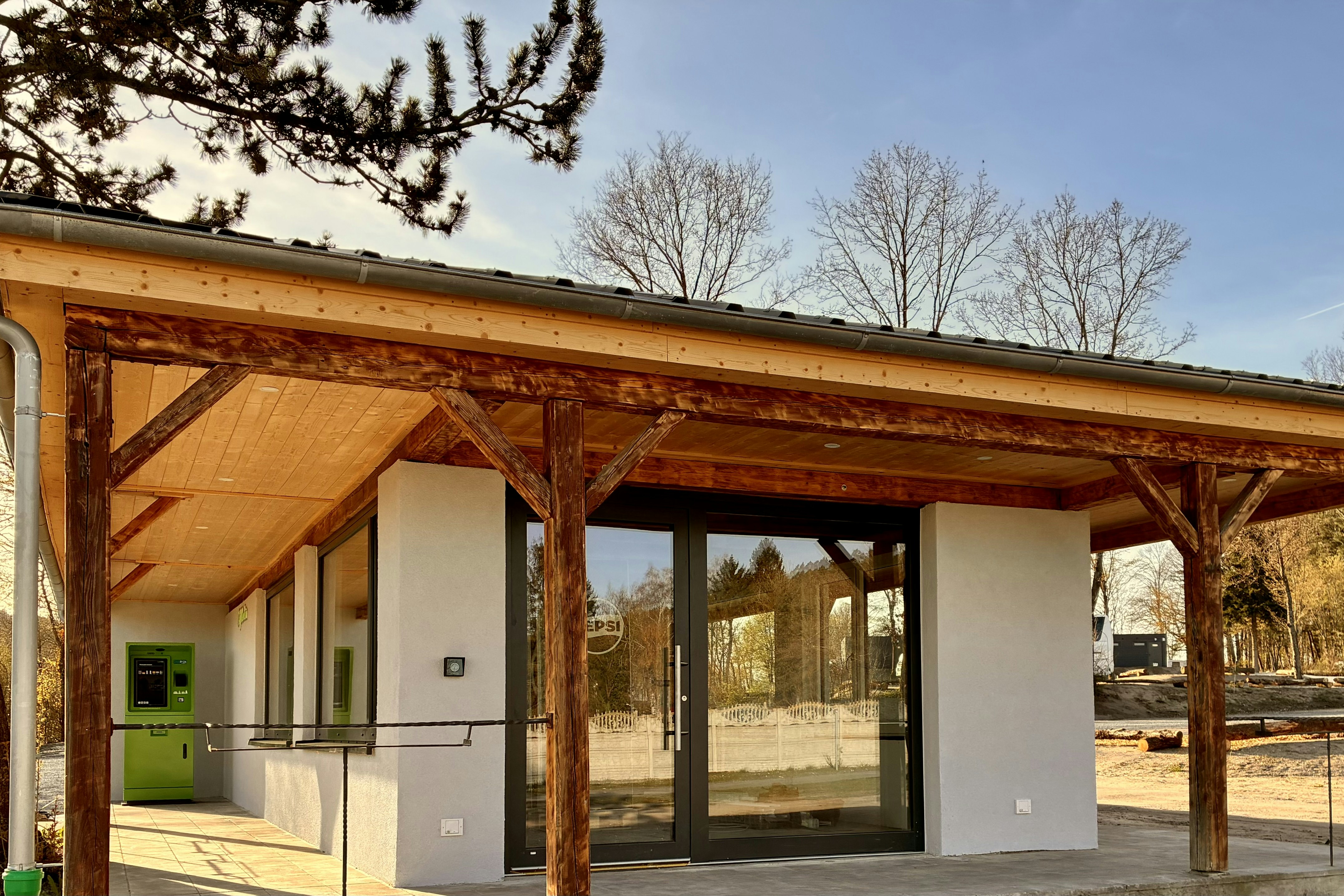 Holzüberdachter Eingang mit Bank und Stützen, daneben weiße Wände und Fenster, im Hintergrund Bäume und ein Weg.