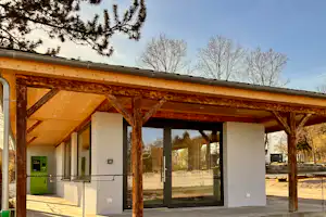 Holzüberdachter Eingang mit Bank und Stützen, daneben weiße Wände und Fenster, im Hintergrund Bäume und ein Weg.