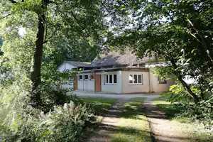 Campinghaus mit Holz- und Putzfassade zwischen Bäumen, Weg zum Eingang und Rasenfläche im Grünen.