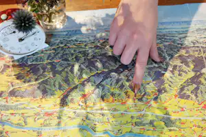 Hand zeigt auf eine Camping-Karte auf dem Tisch, mit Landschaftsdruck und markierten Routen.