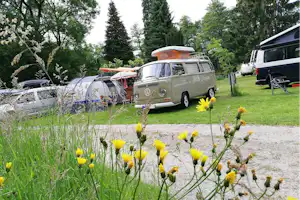 Ein VW-Bus auf dem Campingplatz, daneben ein Wohnmobil und Campingstühle, umgeben von Wiese und Bäumen.