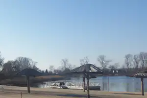 Seeufer mit Sandstrand, Holzpavillons und Stegen am Wasser, im Hintergrund Bäume und Hügel.