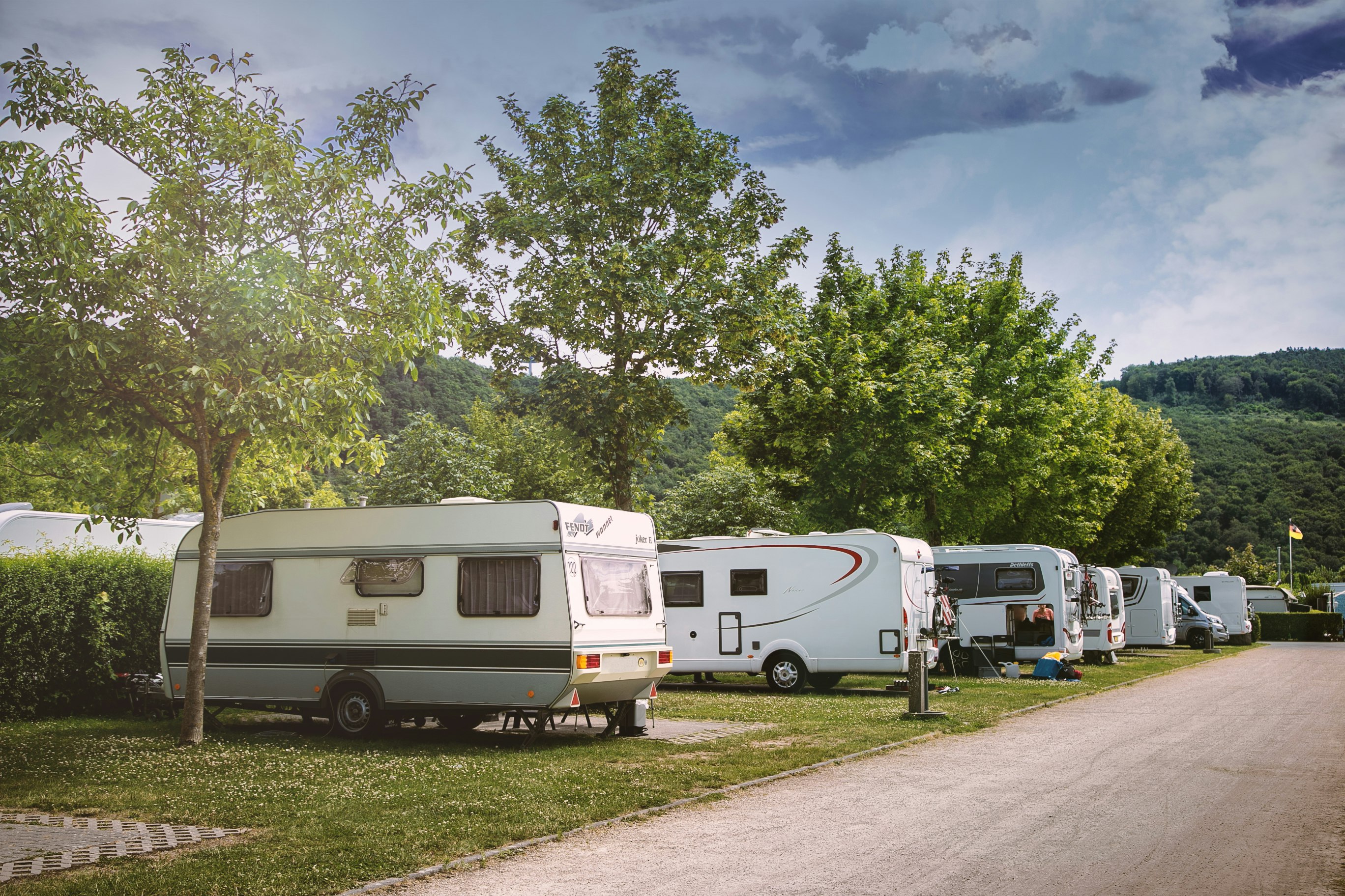 Wohnmobile auf einem Campingplatz entlang eines Weges, mit Bäumen und Hecken im Hintergrund.