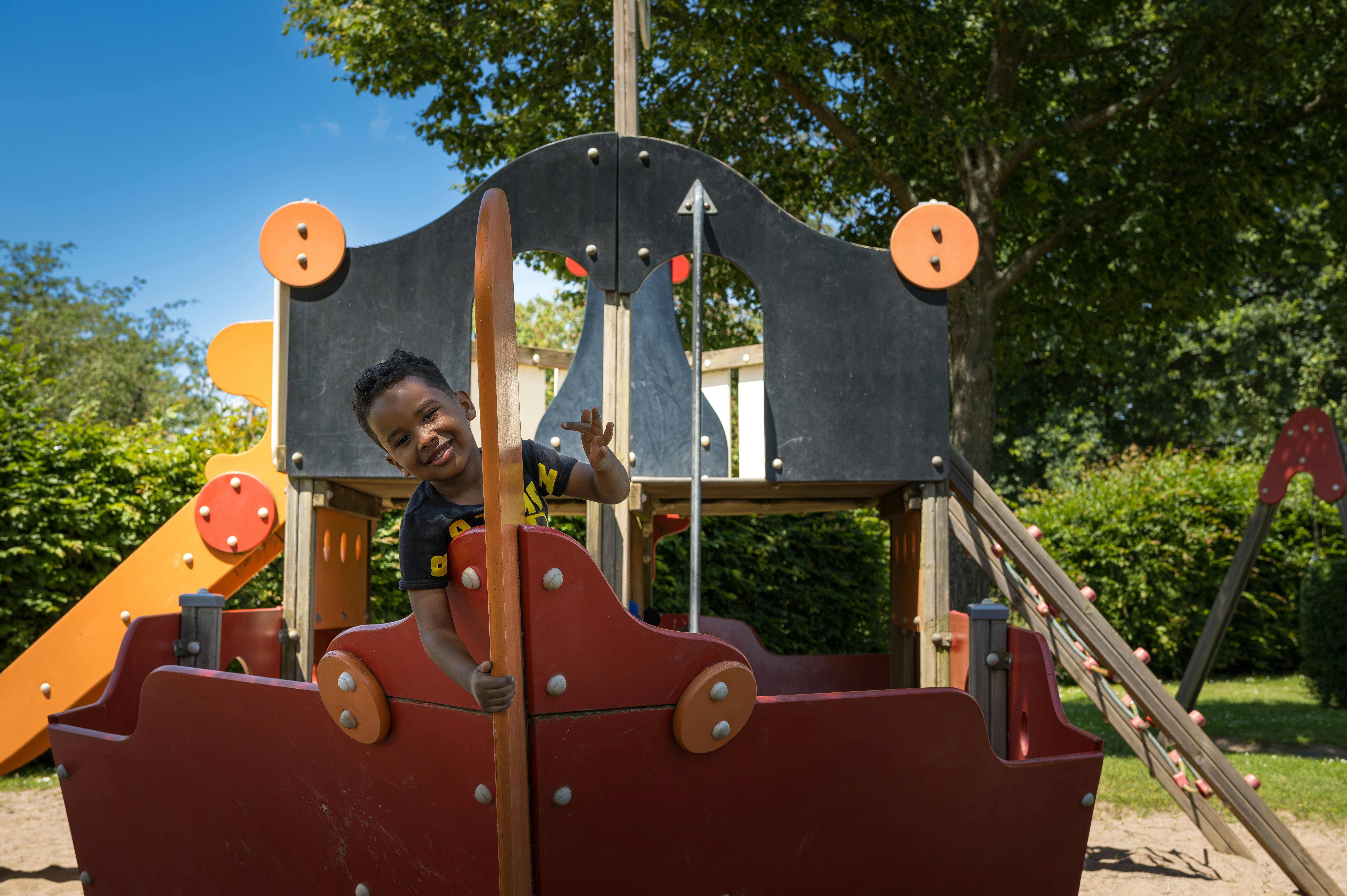Spielplatzschaukel auf dem Campingplatz mit rotem Sitz und Metallgestell, im Hintergrund Bäume und Rasen.