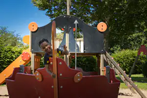 Spielplatzschaukel auf dem Campingplatz mit rotem Sitz und Metallgestell, im Hintergrund Bäume und Rasen.