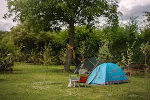 Blaues Campingzelt auf einer Wiese, daneben Campingstühle und Tisch, großer Baum im Hintergrund.