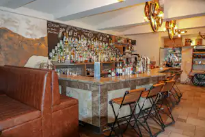 Campingbar mit Theke voller Flaschen, Holzstühlen und Sitzbank im Innenraum, beleuchtet von einer goldenen Lampe.