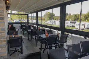 Überdachte Camping-Terrasse mit mehreren Tischen und Stühlen, großen Fenstern und Blick auf Wohnmobile und Stellplätze.
